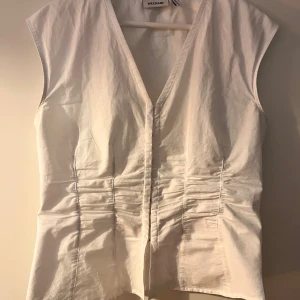 Vit corsett topp från Weekday - Säljer en stilren vit blus från Weekday med v-ringning och insydda detaljer i midjan. Blusen är ärmlös och har en figurnära passform som framhäver siluetten. Perfekt till många olika outfits! Går m-l!