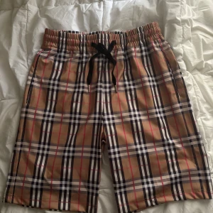 Rutig shorts från Burberry - Snygga rutiga shorts från Burberry i klassiskt beige, svart, vit och röd färgkombination. Modellen har resår i midjan med svart snörning och är tillverkad i 100% bomull. Perfekta för sommaren med sin lätta och luftiga känsla.