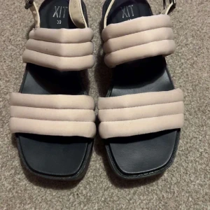 Beige sandaler med vadderade remmar från XIT - Säljer ett par trendiga sandaler från XIT i storlek 39. De har breda, vadderade remmar i beige och en svart, räfflad platt sula. Perfekta för sommaren och ger en cool look till din outfit.