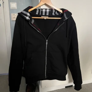 Svart hoodie från Burberry med dragkedja - Svart hoodie från Burberry med klassiskt rutigt foder i huvan och dragkedja framtill. Tröjan har långa ärmar, fickor och justerbar huva med snören. Perfekt för en stilren och bekväm look.