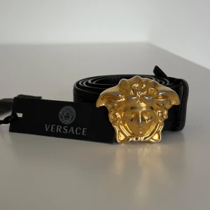 Versace skärp  - Säljer ett svart bälte från Versace i skinn med ett stort, guldfärgat Medusa-spänne. Bältet är tillverkat i Italien och har exklusiv känsla med tydlig Versace-logga. Perfekt accessoar för att lyfta din outfit. Extra hål för att passa en midja ned till cirka 29.
