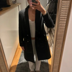 Svart kappa från Carin Wester - Stilren svart kappa från Carin Wester med klassisk krage och dubbelknäppning framtill. Långärmad modell med två fickor och rak passform.