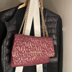 Vinröd axelväska från Valentino (pris diskuteras privat) - Säljer en vinröd axelväska från Valentino med guldfärgad kedja och präglad logotyp över hela väskan. Väskan har ett stort fack och mindre innerfack. Väskan är köpt utomlands därefter €. Väskan är använd ett fåtal gånger, och säljer den för att jag alltid använder en och samma väska❤️PRIS DISKUTERAS, väskan är äkta