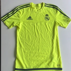 Real Madrid neon gul fotbollströja från Adidas - Säljer en neon gul Real Madrid fotbollströja från Adidas med klubbmärke och Adidas-logga på bröstet. Tröjan har korta ärmar, tunna vertikala ränder och klassiska Adidas-ränder på axlarna. Materialet är lätt och andas, perfekt för träning eller match.