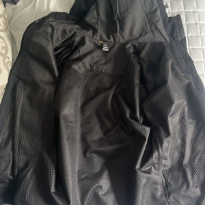 Svart skaljacka från Haglöfs med huva - Säljer en svart skaljacka från Haglöfs med justerbar huva och dragkedja framtill. Jackan har minimalistisk design, meshfoder på insidan och är perfekt för utomhusaktiviteter. Klassisk Haglöfs-logga på bröstet och praktiska fickor.