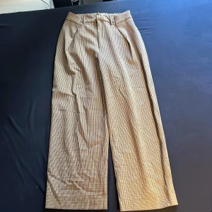 Beige-rutiga kostymbyxor med vida ben - Snygga kostymbyxor i beige och svart med rutig design. Byxorna har hög midja, vida ben och stängs med dragkedja och hake. Perfekta för en dressad look och passar till många olika stilar.