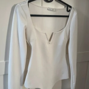 Vit långärmad bodysuit från NLY One - Säljer en stilren vit bodysuit från NLY One med lång ärm och djup v-ringning framtill. Toppen har en figurnära passform och är tillverkad i ett stretchigt material som sitter snyggt på kroppen. Perfekt att styla till jeans eller kjol.