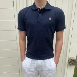 Mörkblå pikétröja från Ralph Lauren - Säljer en klassisk mörkblå pikétröja från Ralph Lauren med vit broderad logga på bröstet. Tröjan har korta ärmar, krage och knappar framtill. Perfekt för en stilren och avslappnad look. Den sitter riktigt bra, exakt som en piké ska sitta!! Den är s men passar men skulle säga att den passar mer som M