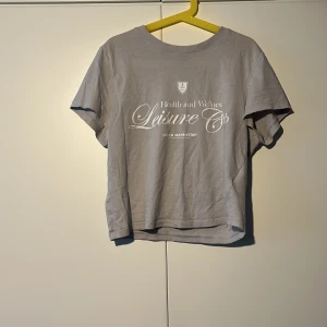 Ljusgrå t-shirt från Divided - Ljusgrå t-shirt från Divided med vit text framtill: 'Health and Wellness Leisure Ops, Lower Manhattan'. Klassisk rund hals och korta ärmar. Tillverkad i mjuk bomull för en skön känsla.