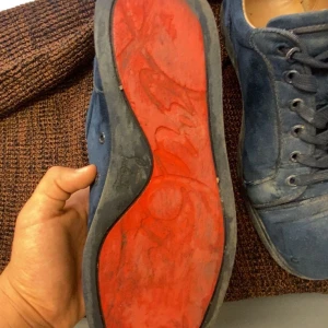 Blå sneakers med röd sula - Säljer ett par blå sneakers med snörning och röd sula. Skorna har rund tå och platt sula, samt är tillverkade i mocka. Perfekta för dig som gillar en stilren och avslappnad look.