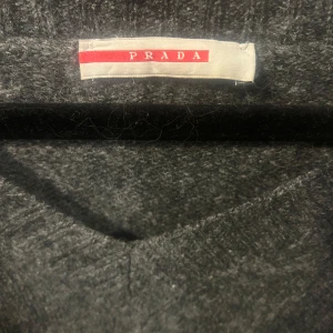 Mörkgrå v-ringad tröja från Prada - Mörkgrå v-ringad tröja från Prada i mjukt material. Klassisk och stilren design med långa ärmar och ribbade muddar. Perfekt för dig som gillar en enkel men lyxig look. Mycket bra skick