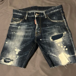 Avklippt jeansshorts från Dsquared2 - Riktigt snygga mörkblå avklippt jeansshorts från Dsquared2 med slitningar, hål och vit färgstänk. Klassisk femficksmodell med råa kanter och tryck på bakfickan. Perfekta för dig som gillar streetwear och vill sticka ut.