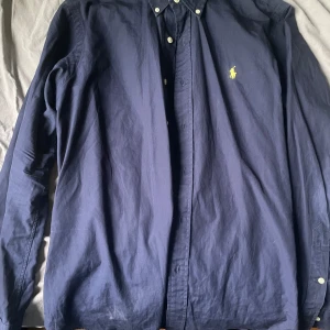 Mörkblå skjorta från Ralph Lauren - Säljer en klassisk mörkblå skjorta från Ralph Lauren med gul broderad logga på bröstet. Skjortan har lång ärm, knappar hela vägen och button-down krage. Perfekt för dig som gillar stilrena plagg med snygg passform.