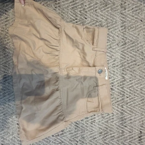 Beige kortkjol med volang - Snygg beige kortkjol med volangdetalj och klassisk jeansknäppning. Kjolen har fickor fram och bälteshällor i midjan. Perfekt för en trendig och avslappnad look.