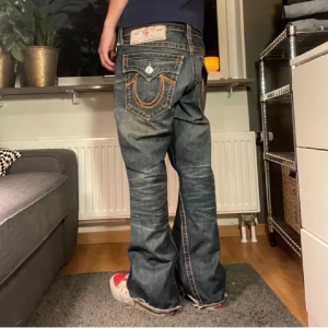 true religion jeans - Säljer ett par blå baggy jeans med tydliga kontrastsömmar och stora bakfickor med dekorativa detaljer. Jeansen har normal passform i midjan och vida ben. Perfekta för en avslappnad streetstil.