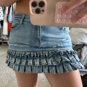 Jeanskjol med volanger - Supersöt kort jeanskjol med två lager volanger nedtill. Klassisk blå denim med fickor fram och bak samt dragkedja och knapp i midjan. Perfekt för dig som vill sticka ut lite extra!