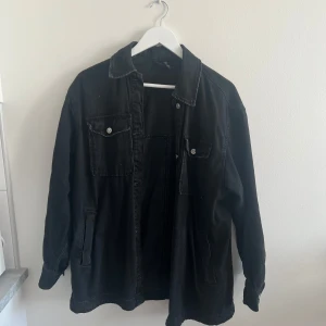 Svart jeansjacka med knappar - Säljer en svart jeansjacka med klassisk krage och knäppning framtill. Jackan har två bröstfickor med lock och knappar samt två sidofickor. Perfekt för dig som gillar en enkel och stilren look.