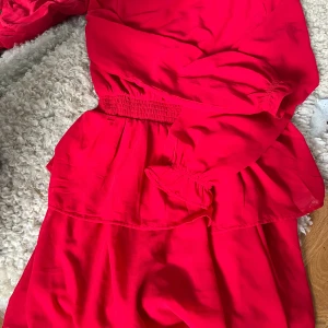 Röd klänning med volang - Säljer en röd klänning med volangdetaljer och resår i midjan. Klänningen är ärmlös och har en luftig, lätt form som passar perfekt till sommarens tillfällen.