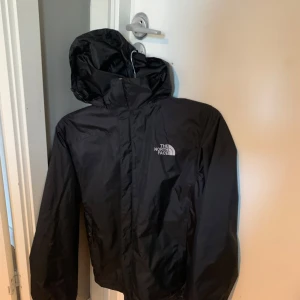 Svart vindjacka från The North Face - Säljer en svart vindjacka från The North Face med huva och dragkedja framtill. Jackan har en stilren design med logga på bröstet och är perfekt för blåsiga dagar. Passar både till promenader och utomhusaktiviteter.