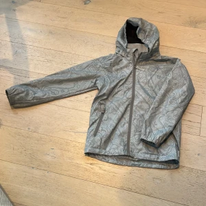 Grå mönstrad tunn jacka från Polarn O. Pyret - Säljer en grå tunn jacka från Polarn O. Pyret med abstrakt mönster och huva. Jackan har dragkedja framtill och två fickor med dragkedja. Perfekt för blöta dagar och håller dig torr med stil. Den är även vattenavvisande.