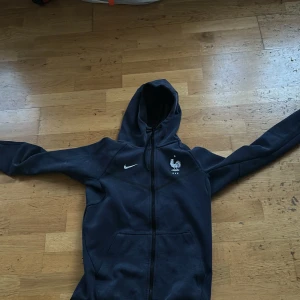 Mörkblå Nike hoodie med Frankrikes fotbollslandslag - Säljer en mörkblå hoodie från Nike med Frankrikes fotbollslandslags logga på bröstet. Jackan har huva, dragkedja och två fickor framtill. Perfekt för dig som gillar fotboll och vill ha en sportig stil.