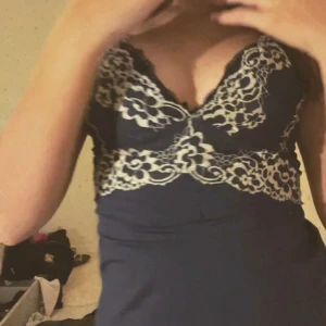 Mörkblå klänning med spetsdetaljer - Säljer en mörkblå klänning med v-ringning och dekorativa vita spetsdetaljer över byst och midja. Klänningen har en figurnära passform och är ärmlös, perfekt för en elegant look.