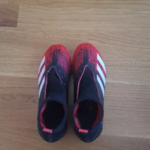 Adidas fotbollsskor i svart och rött - Säljer ett par Adidas fotbollsskor i svart och rött med vita detaljer. Skorna har en strumpliknande passform utan traditionell snörning och är designade för bra grepp på planen med fasta dobbar undertill. Pris kan diskuteras