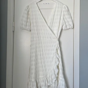 Vit omlottklänning med volang från MNG - Säljer en söt vit omlottklänning från Mango med korta puffärmar och volangdetalj längs nederkanten. Klänningen har knyt i midjan och ett diskret rutmönster i tyget. Perfekt för sommarens tillfällen!