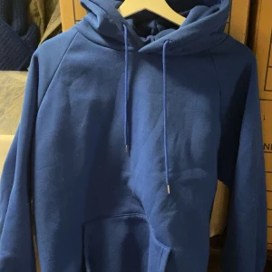 Blå hoodie med huva och ficka - Säljer en enkel blå hoodie med huva och dragsnören. Tröjan har en stor magficka och långa ärmar. Perfekt för en avslappnad stil eller när du vill vara extra bekväm.
