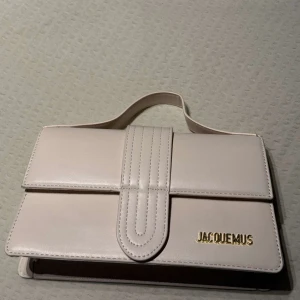 Vit handväska från Jacquemus - Säljer en stilren vit handväska från Jacquemus med guldfärgad logga framtill. Väskan har ett elegant handtag och axelremsband tillkommer💕