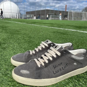 Svarta sneakers från Saint Laurent - Säljer ett par svarta sneakers från Saint Laurent med vit sula och vita snören. Skorna har en sliten look med synliga slitningar och Saint Laurent-logga på sidan. Perfekta för dig som gillar en avslappnad och trendig stil.