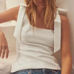 Vit linnetopp med rosettband - Snygg vit linnetopp med breda axelband som knyts i rosetter. Toppen har en rak, något figurnära passform och är perfekt att matcha med jeans eller kjol för en trendig look. Kan diskutera pris vid snabbt köp