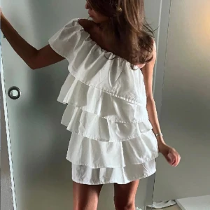 Vit volangklänning med one shoulder - Intressekoll på denna fina vit kortklänning med flera volanglager och one shoulder-design. Klänningen har en luftig och lekfull stil, perfekt för varma dagar eller speciella tillfällen. Den är axelbandslös på ena sidan och har en snygg, asymmetrisk form.