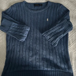 Blå kabelstickad tröja från Polo Ralph Lauren - Kabelstickad blå tröja från Polo Ralph Lauren med rund halsringning och broderad logga på bröstet. Tröjan har långa ärmar och ribbade muddar vid ärmslut och nederkant. Perfekt för lager-på-lager och klassisk stil.