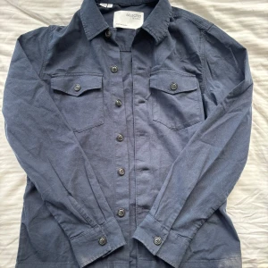 Mörkblå overshirt från Selected Homme - Snygg mörkblå overshirt från Selected Homme med två bröstfickor och knappar framtill. Skjortan har klassisk krage och långa ärmar med knappar vid ärmsluten. Perfekt för en stilren look.