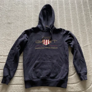 Mörkblå GANT hoodie ”American Sportswear” - Säljer en mörkblå hoodie från GANT med broderad logga och sköld på bröstet. Tröjan har huva med dragsko och en stor magficka. Klassisk och stilren design med guldfärgade detaljer och texten 'American Sportswear'.