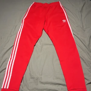Röda Adidas mjukisbyxor med vita ränder - Säljer ett par klassiska röda Adidas mjukisbyxor med vita ränder längs sidorna och elastisk midja. Byxorna har en smal passform och är perfekta för en sportig och avslappnad stil. Adidas-logga på ena benet.
