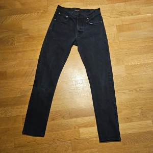 Svarta jeansbyxor från Nudie Jeans w28 L 32 - Säljer ett par svarta jeans från Nudie Jeans med klassisk femficksdesign och raka ben. Jeansen har normal passform och är tillverkade i denim med snygga detaljer vid fickorna. Perfekta för dig som gillar stilrena och tidlösa jeans. Inga returer