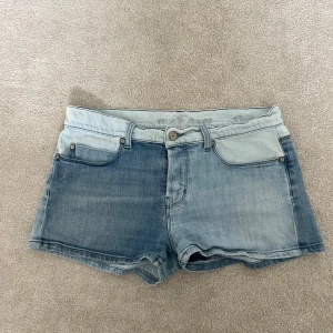 Trefärgade jeansshorts från MHI Jeans - Säljer ett par coola tvåfärgade jeansshorts från MHI Jeans. Shortsen har klassisk femficksdesign, låg midja och är sydda i två olika blå denimnyanser. Perfekta för sommaren och ger en unik look. Storlek 27.