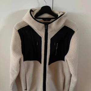Vit fleecejacka med svarta detaljer från Sail Racing - Säljer en vit fleecejacka med svarta partier och dragkedjor från Sail Racing. Jackan har huva, hel dragkedja framtill och två bröstfickor med dragkedja. Perfekt för kyliga dagar och snygg att ha lager på lager.