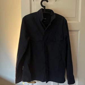 Mörkblå skjorta från Only & Sons - Säljer en stilren mörkblå skjorta från Only & Sons. Skjortan har långa ärmar, klassisk krage och knäppning framtill. Två bröstfickor med lock ger en snygg detalj. Perfekt för dig som gillar enkel och clean stil.