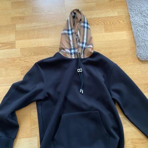 Burberry - Säljer en svart hoodie från Burberry med klassiskt rutmönstrad huva i beige, svart, vit och blå. Tröjan har en stor ficka framtill och justerbara snören med B-detalj vid huvan. Perfekt för en trendig och avslappnad stil. Passar S/M/L