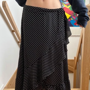 Svart prickig omlottkjol - Svart långkjol med vita prickar och volangkant. Kjolen har omlottmodell och knyts i midjan för en snygg passform. Perfekt för dig som gillar en flowy och trendig stil. 