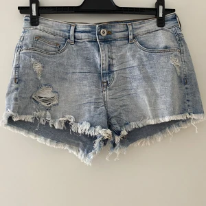 Ljusblå jeansshorts med slitningar och fransar - Säljer ett par ljusblå jeansshorts med slitna detaljer och fransig kant. Shortsen har klassisk femficksmodell, knapp och dragkedja framtill samt råa kanter nedtill. Perfekta för sommaren!