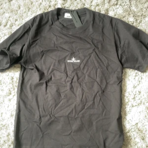 Svart t-shirt från Stone Island - Säljer en svart t-shirt från Stone Island med vit logga på bröstet och stor grafisk logga på ryggen. Klassisk rund hals och korta ärmar. Perfekt för dig som gillar streetwear och vill ha något stilrent med tydlig branding.