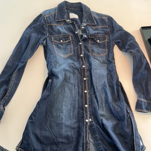 Jeansskjorta med bälte från Peak Performance - Säljer en snygg jeanskläning från Peak Performance i mörkblå denim. Kläningen har långa ärmar, två bröstfickor med knappar och ett matchande bälte i midjan. Storlek S (dam). Nypris: ca 1500 kr och mitt: 460 kr. Perfekt för en cool och avslappnad stil. Skriv bara om du har frågor, priset kan diskuteras!