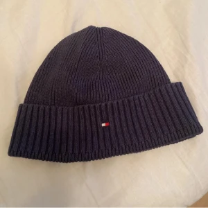 Tommy Hilfiger Mössa - Säljer en mörkblå ribbstickad mössa från Tommy Hilfiger med klassisk uppvikt kant och liten broderad logga framtill. Perfekt för kyliga dagar och enkel att matcha med olika outfits.