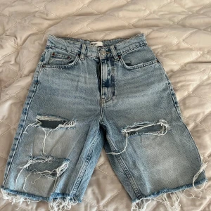 Ljusblå slitna jeansshorts från Ginatricot - Säljer ett par ljusblå jeansshorts från Ginatricot med råa kanter och stora slitningar framtill. Klassisk femficksmodell med hög midja och dragkedja. Perfekta för en avslappnad stil.