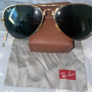 Ray-Ban Aviator solglasögon med guldfärgade bågar - Klassiska Ray-Ban Aviator solglasögon med tunna guldfärgade metallbågar och mörkgröna glas. Ikonisk pilotform och Ray-Ban-logga på linsen. Kommer med originalfodral och putsduk. Går att vika på mitten i en kompakt form.