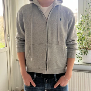 Grå hoodie från Polo Ralph Lauren (M) - Säljer en klassisk grå hoodie från Polo Ralph Lauren med dragkedja och huva. Tröjan har ribbade muddar, två fickor fram och den ikoniska broderade loggan på bröstet. Perfekt för en avslappnad stil.